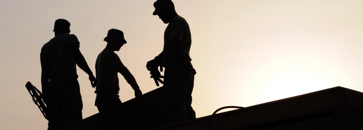Black silhouettes of three blue collar men working on a roof with the sun going down behind them